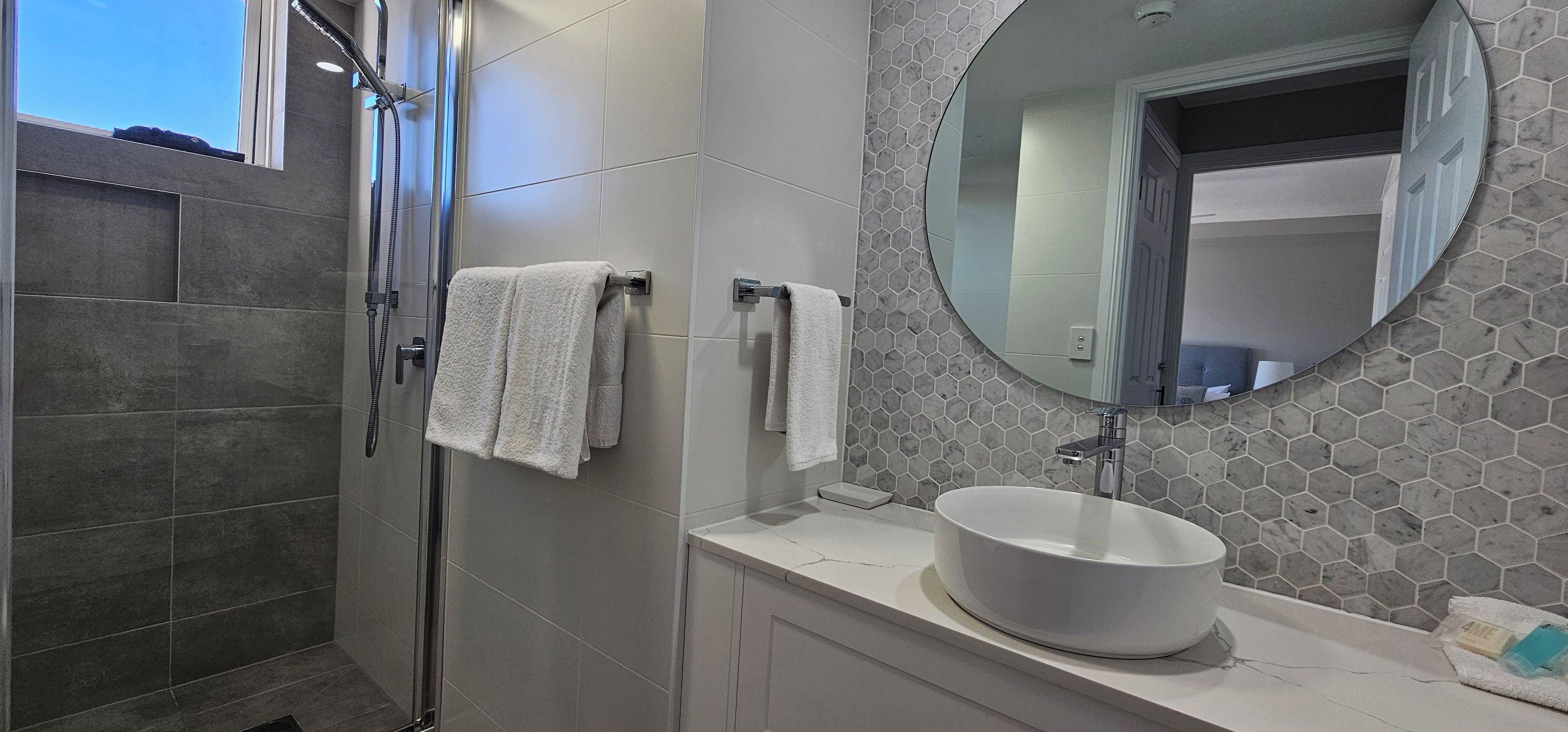 A modern bathroom with a glass-enclosed shower, a round mirror, and a white sink.