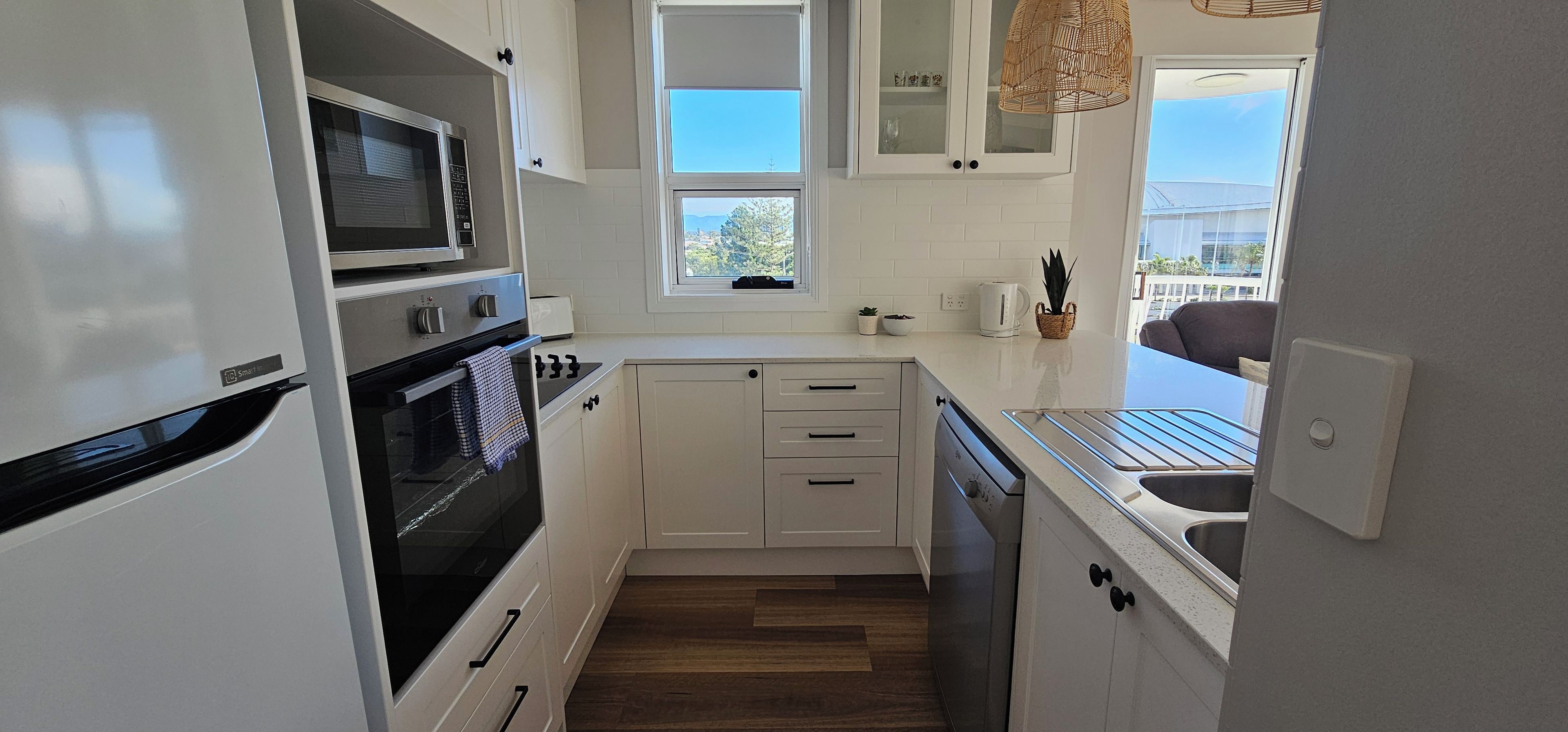 Cocina compacta con muebles blancos, microondas integrado, horno y lavavajillas. Una ventana proporciona luz natural y vistas al exterior.