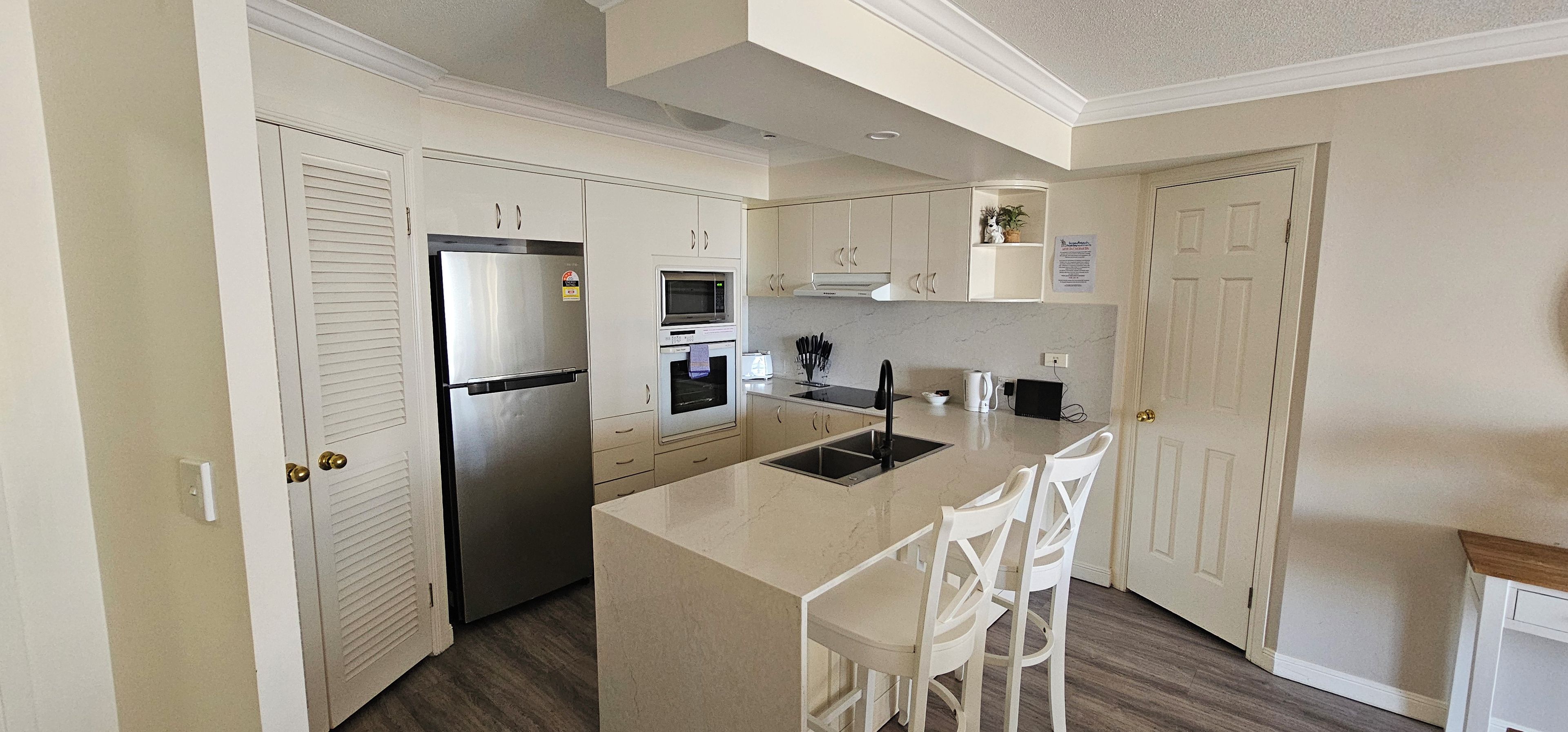 A kitchen with white cabinets, a stainless steel refrigerator, an oven, a microwave, and a sink. There are two white chairs at the island.