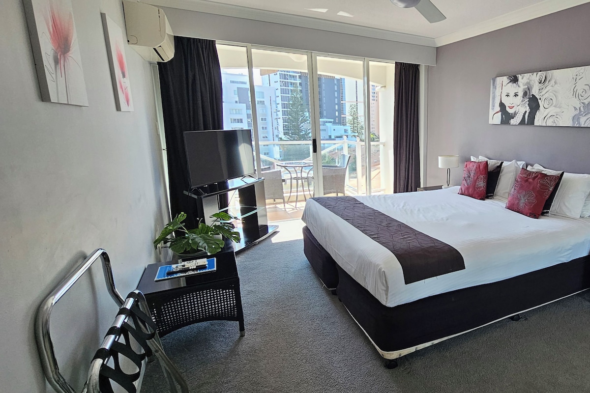 Una habitación de hotel moderna con una cama grande, un escritorio con una computadora, una silla y un balcón con vistas a la ciudad.