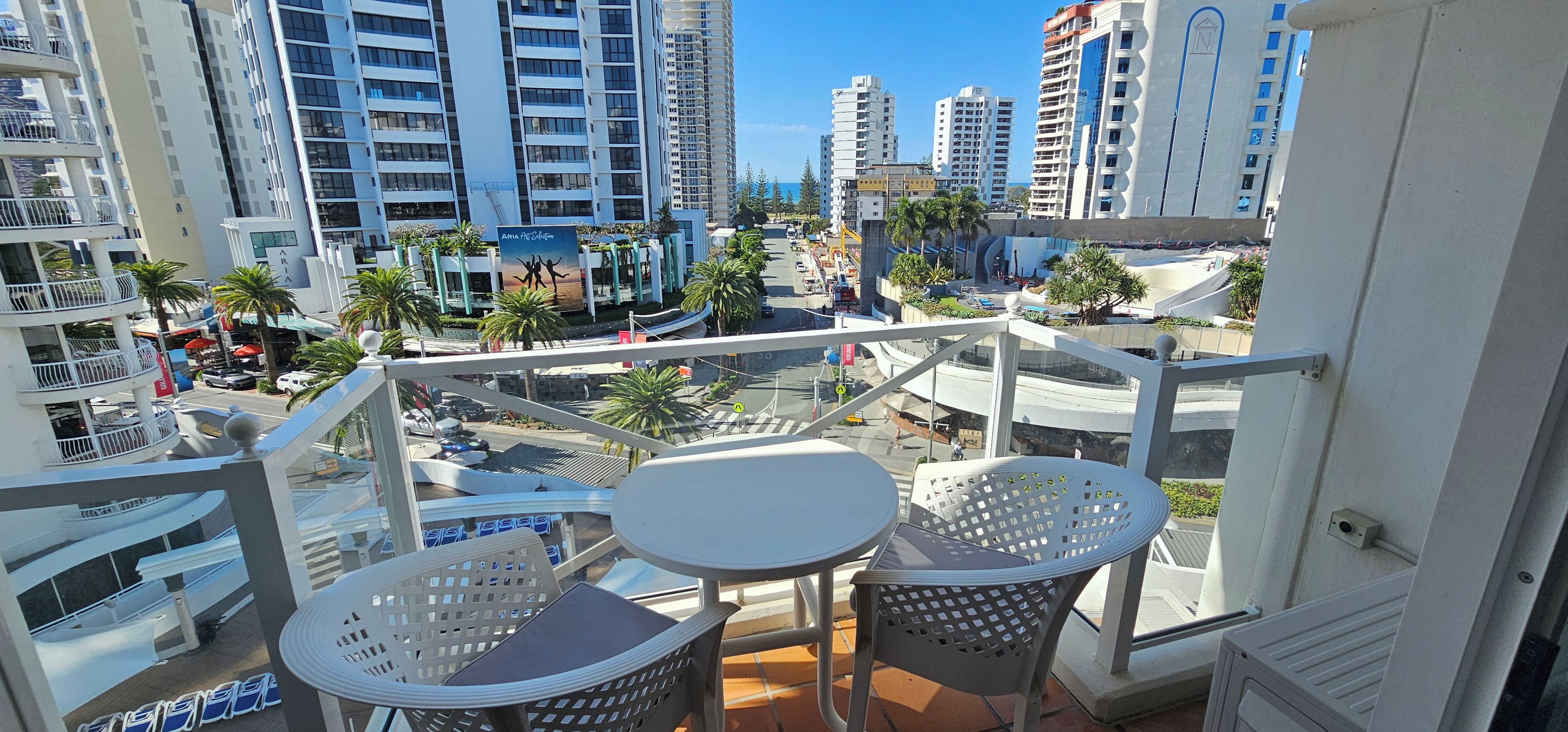 A balcony with two white tables and chairs, overlooking a cityscape with tall buildings and palm trees.
