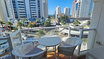 A balcony with two white tables and chairs, overlooking a cityscape with tall buildings and palm trees.