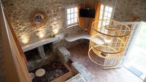 Una habitación de paredes de piedra con un banco de piedra, una lámpara de techo de mimbre y un ventanal con cortinas.