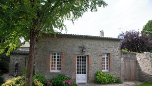 Una casa de piedra con una puerta blanca, dos ventanas con contraventanas y un pequeño jardín con flores rosadas.