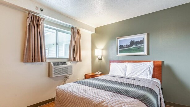 A hotel room with a bed, a window with curtains, an air conditioning unit, a bedside lamp, and a framed picture on the wall. - Sun Suites of Louisville (Louisville)