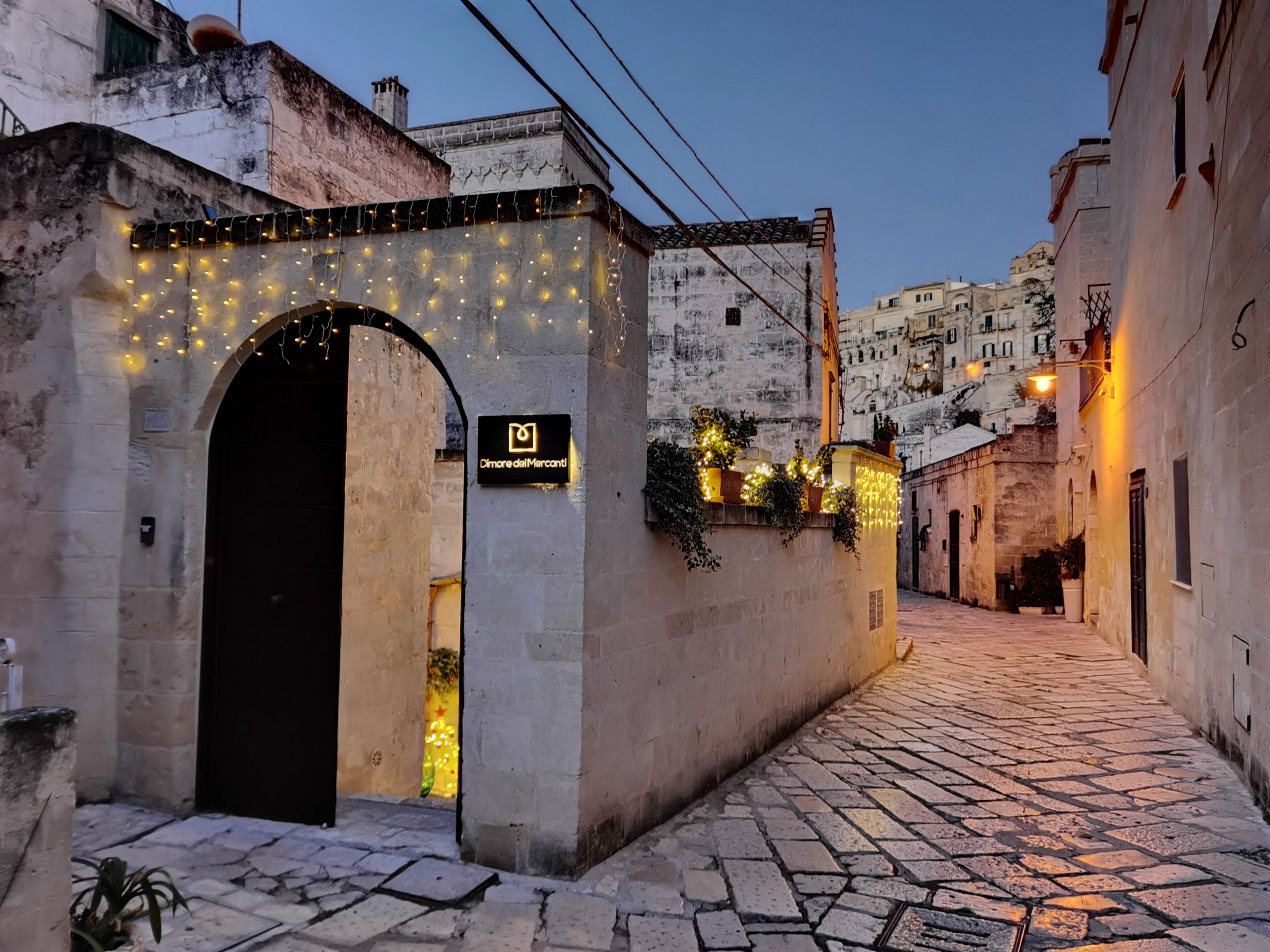 Une ruelle étroite pavée, bordée d’un bâtiment doté d’une entrée en arc de cercle et illuminée de guirlandes lumineuses.