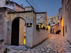 A narrow cobblestone street with a building featuring an arched entrance and string lights. - Dimore dei Mercanti (Matera)