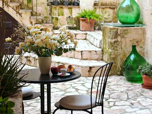Un patio con suelo de piedra, una mesa, sillas, un jarrón de flores, una botella verde y una planta en maceta.