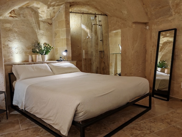 A bedroom with a large bed, a nightstand, a mirror, and a shower area. - Dimore dei Mercanti (Matera)