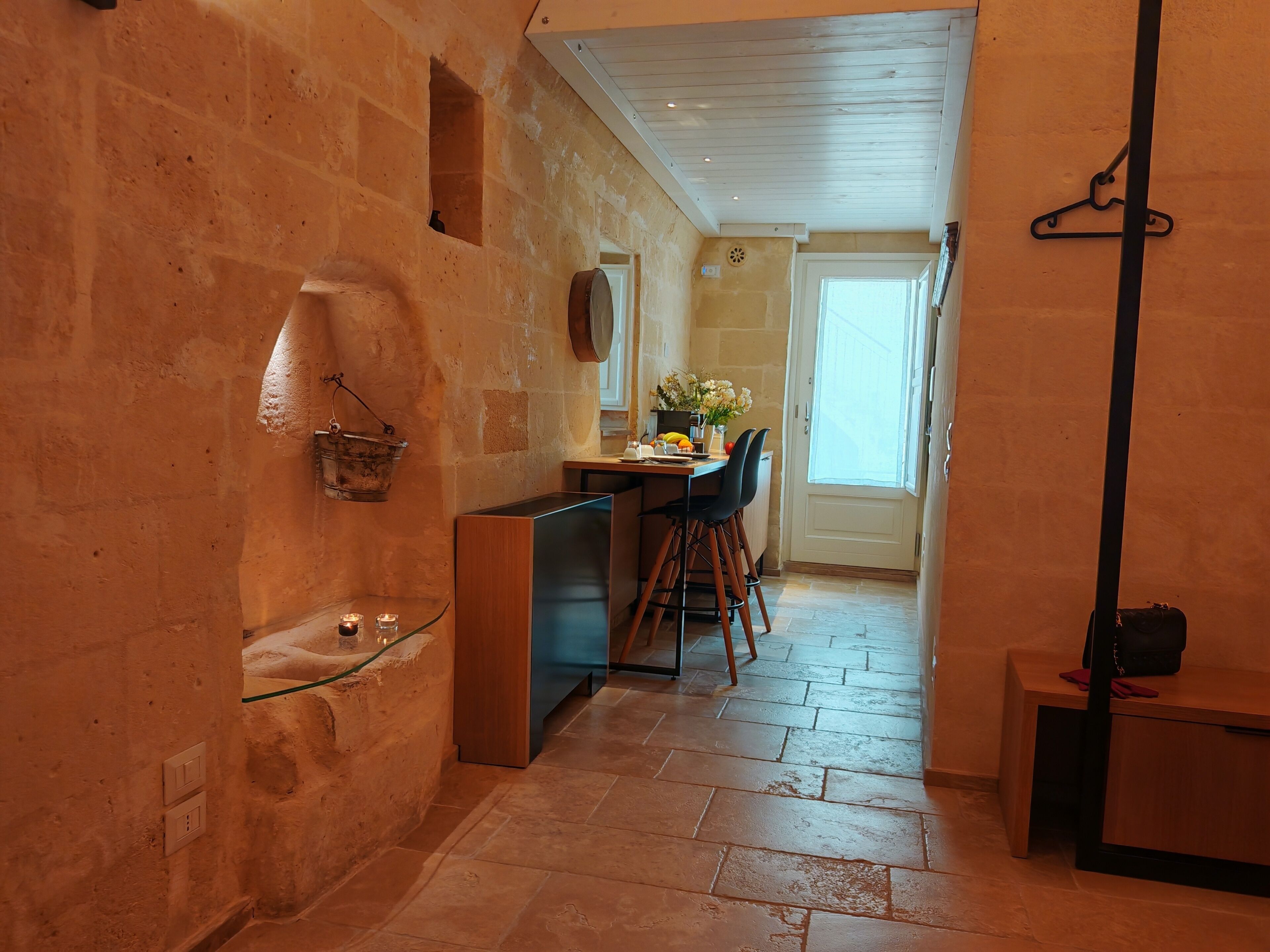 A narrow hallway with stone walls, a small kitchen area with a sink, a dining table with chairs, and a door leading to another room.