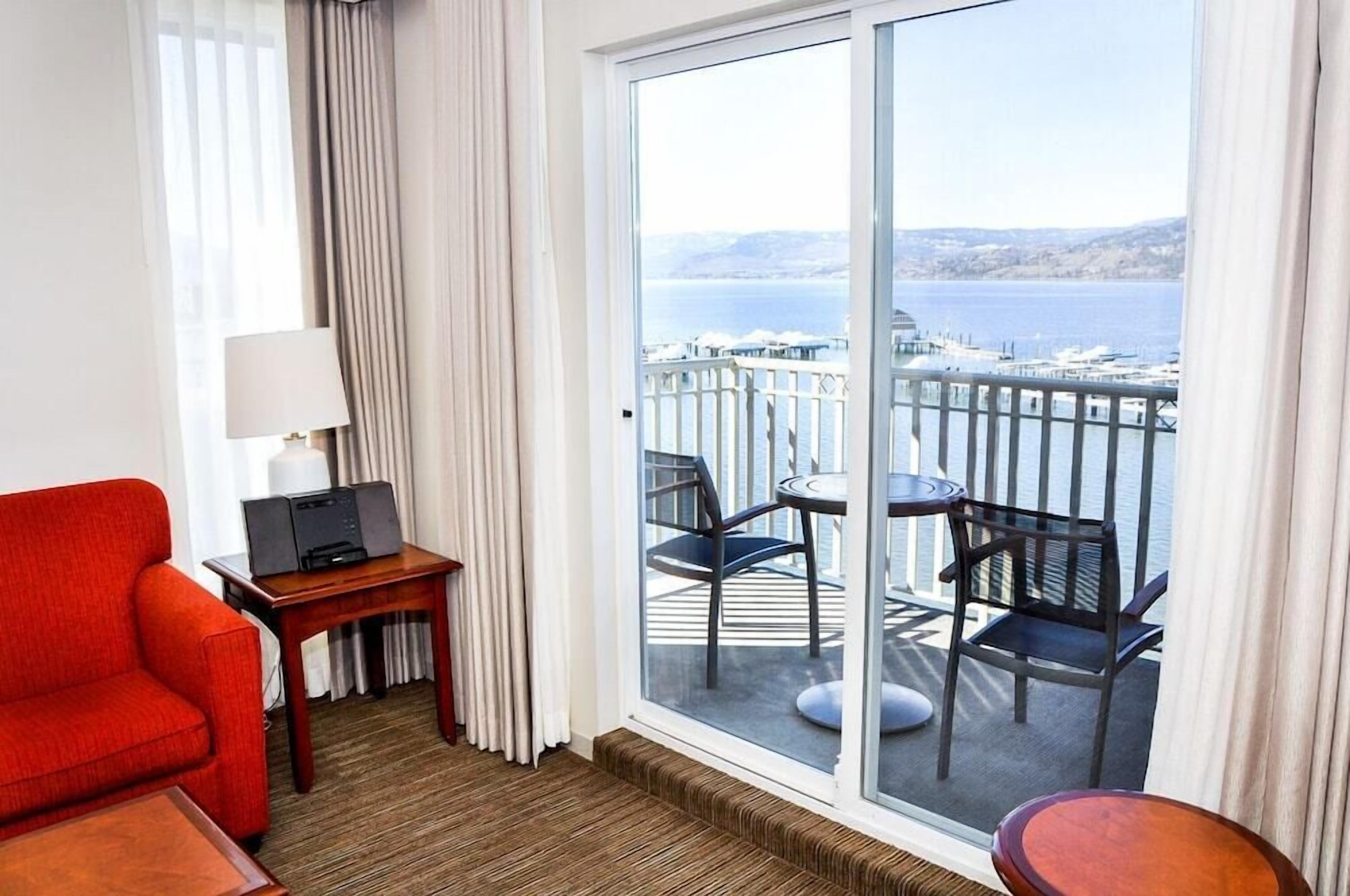 A hotel room with a balcony, a red armchair, a small table with a lamp, and a view of the sea and boats.