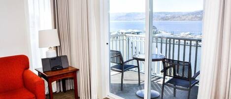 A hotel room with a balcony, a red armchair, a small table with a lamp, and a view of the sea and boats.