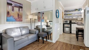 A modern living room with a grey leather sofa, a glass door leading to a kitchen, and a large abstract painting on the wall. - Bayfront Studio, Walk to Beach & Shops | Edgewater (Newport Beach)