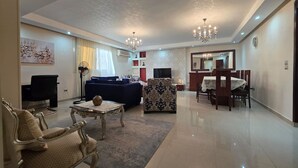 A spacious living room with a blue sofa, a patterned armchair, and a dining area with a table and chairs. - Executive Exclusive Cairo Mohandessin Giza Apart. (Gazirat Mit Oqbah)