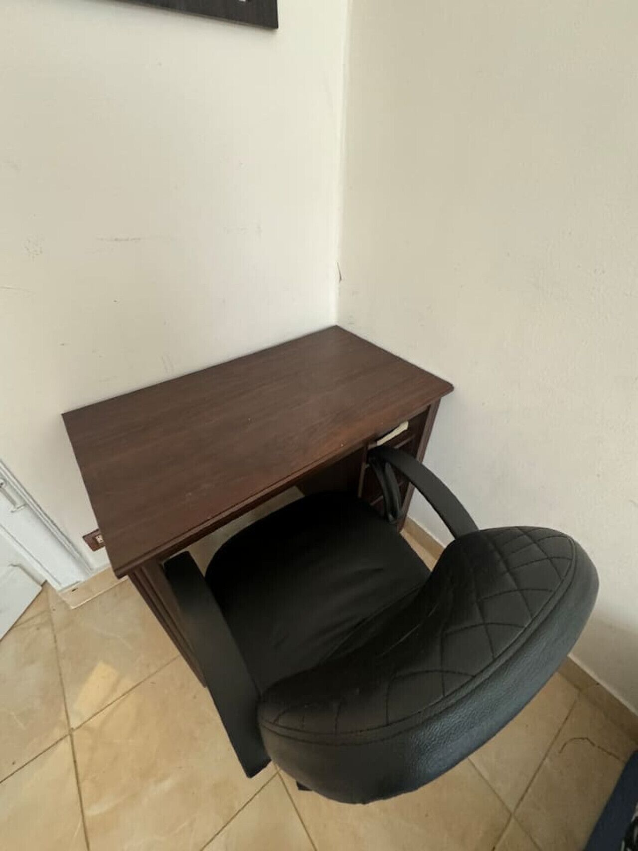 A small, dark wooden desk with a black office chair in a corner of a room with white walls and tiled flooring.