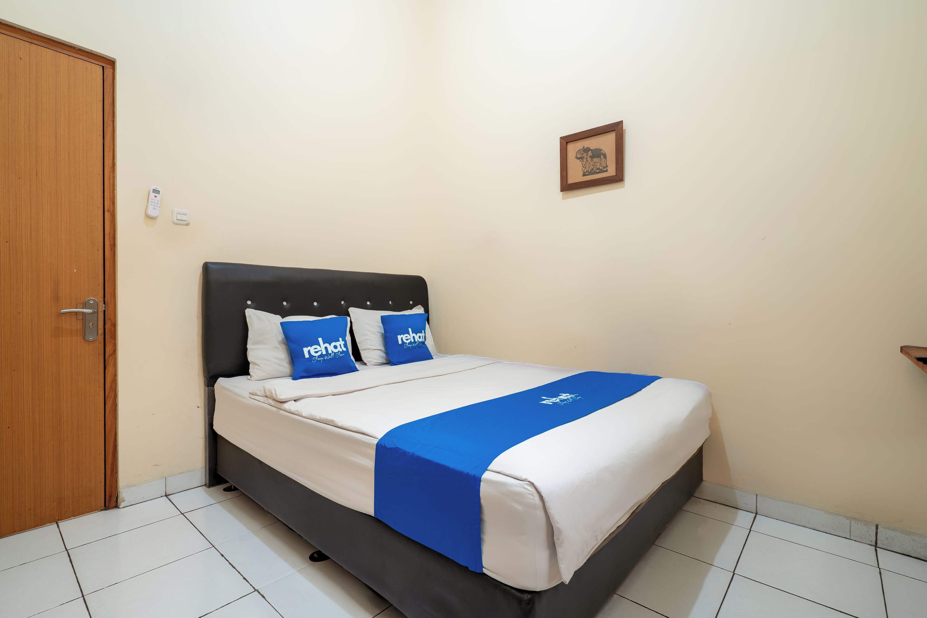 A neatly made bed with a blue and white bedspread, two pillows with 'rehat' branding, and a wooden door to the left.