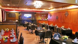 Un restaurant amb diverses taules preparades per dinar, una zona de bar i una pantalla de televisió al fons.