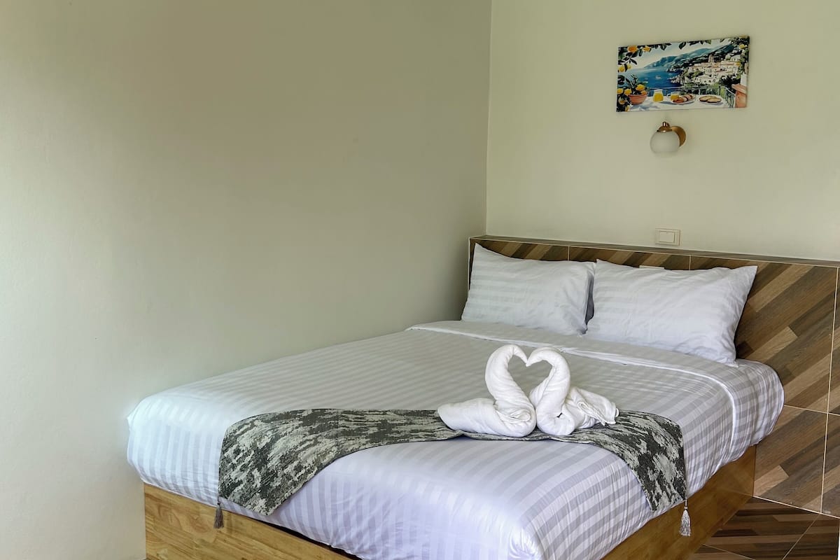 A neatly made bed with a heart-shaped towel, a wall-mounted air conditioner, and a painting of a coastal scene.