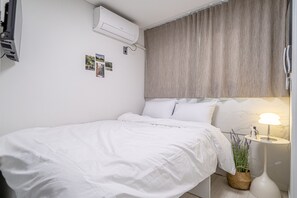 En pænt redt seng med hvide sengetøj, en vægmonteret klimaanlæg, et lille natbord med en lampe og en vægmonteret telefon.