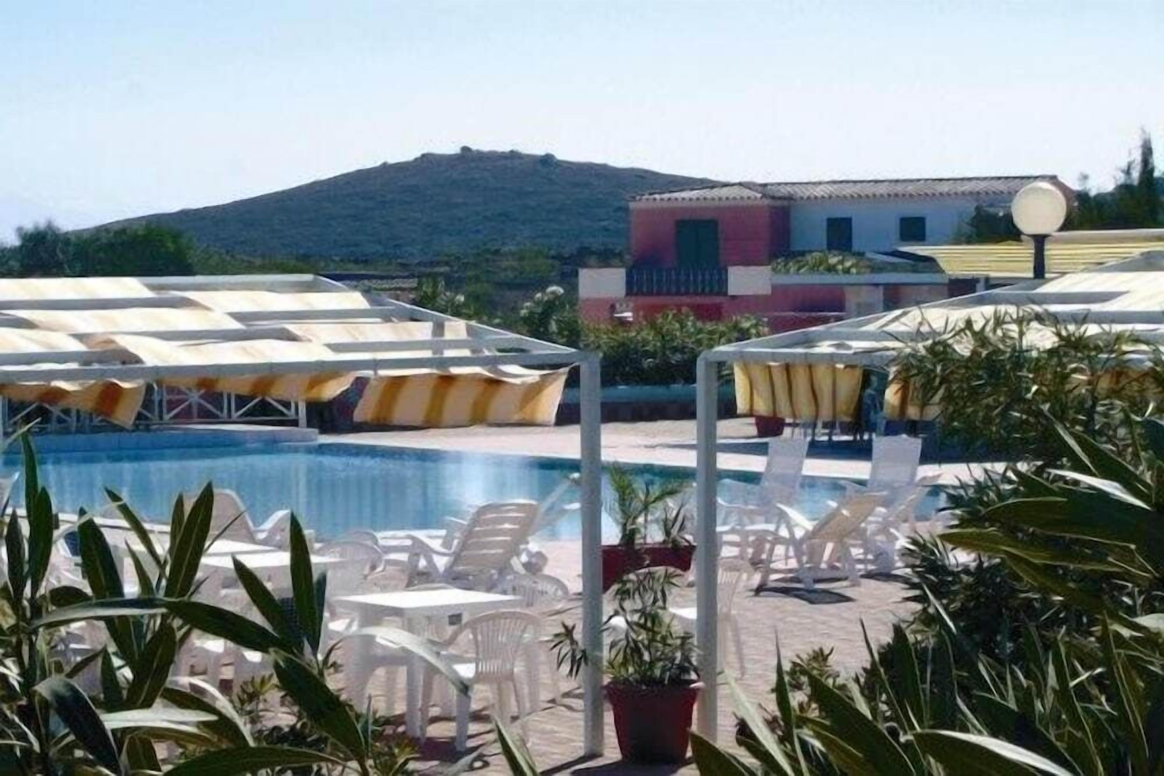 A pool area with sun loungers and umbrellas, surrounded by potted plants and a mountain in the background.