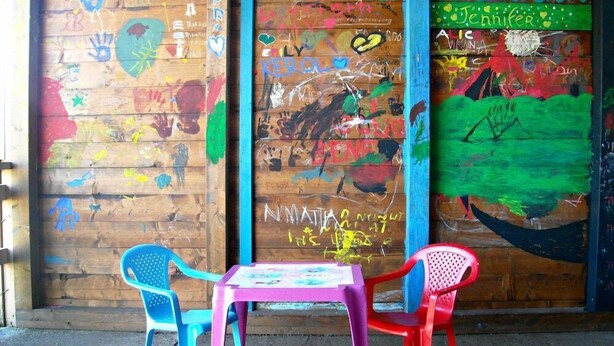 Paret de fusta amb grafitis i petjades de mans de colors. Hi ha dues cadires de plàstic i una taula petita al davant.