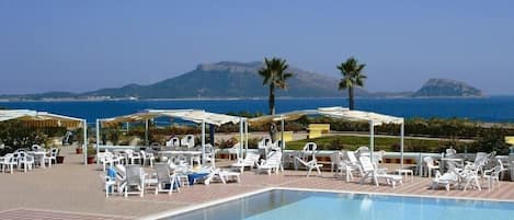 A pool area with lounge chairs and umbrellas, a clear view of the sea, and a mountain in the background.