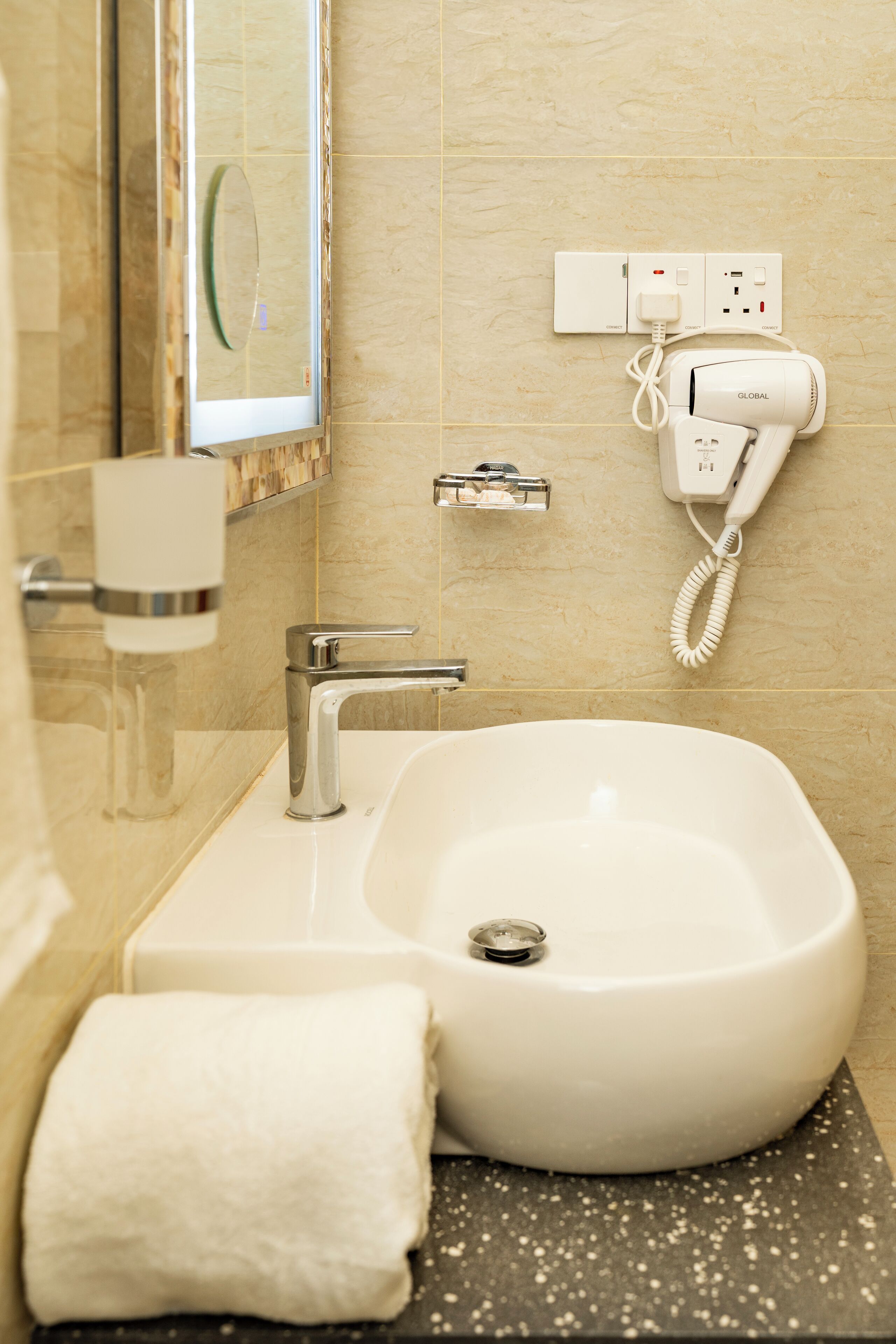 Un baño con lavamanos blanco, espejo, toalla y secador de pelo enchufado a un enchufe.