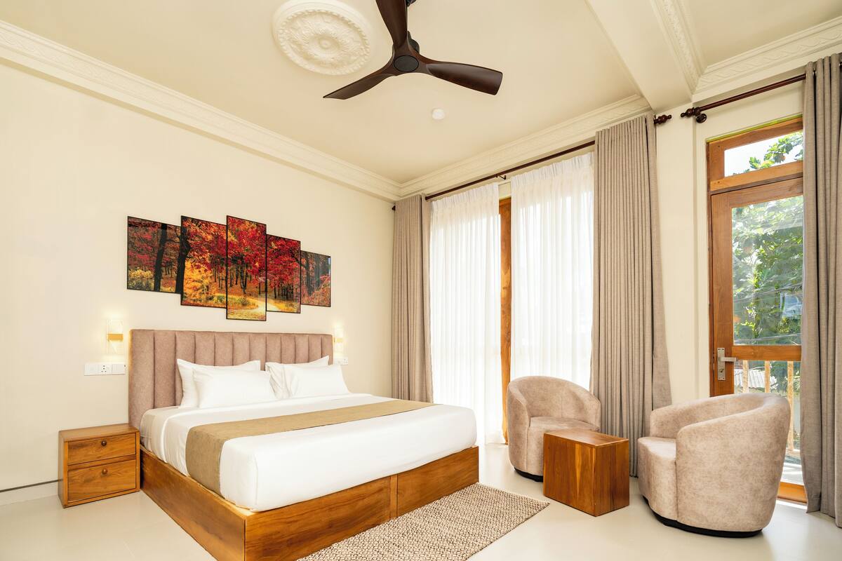 Habitación de hotel moderna con una cama grande, un ventilador de techo, un área de descanso con dos sillones y un ventanal con cortinas.