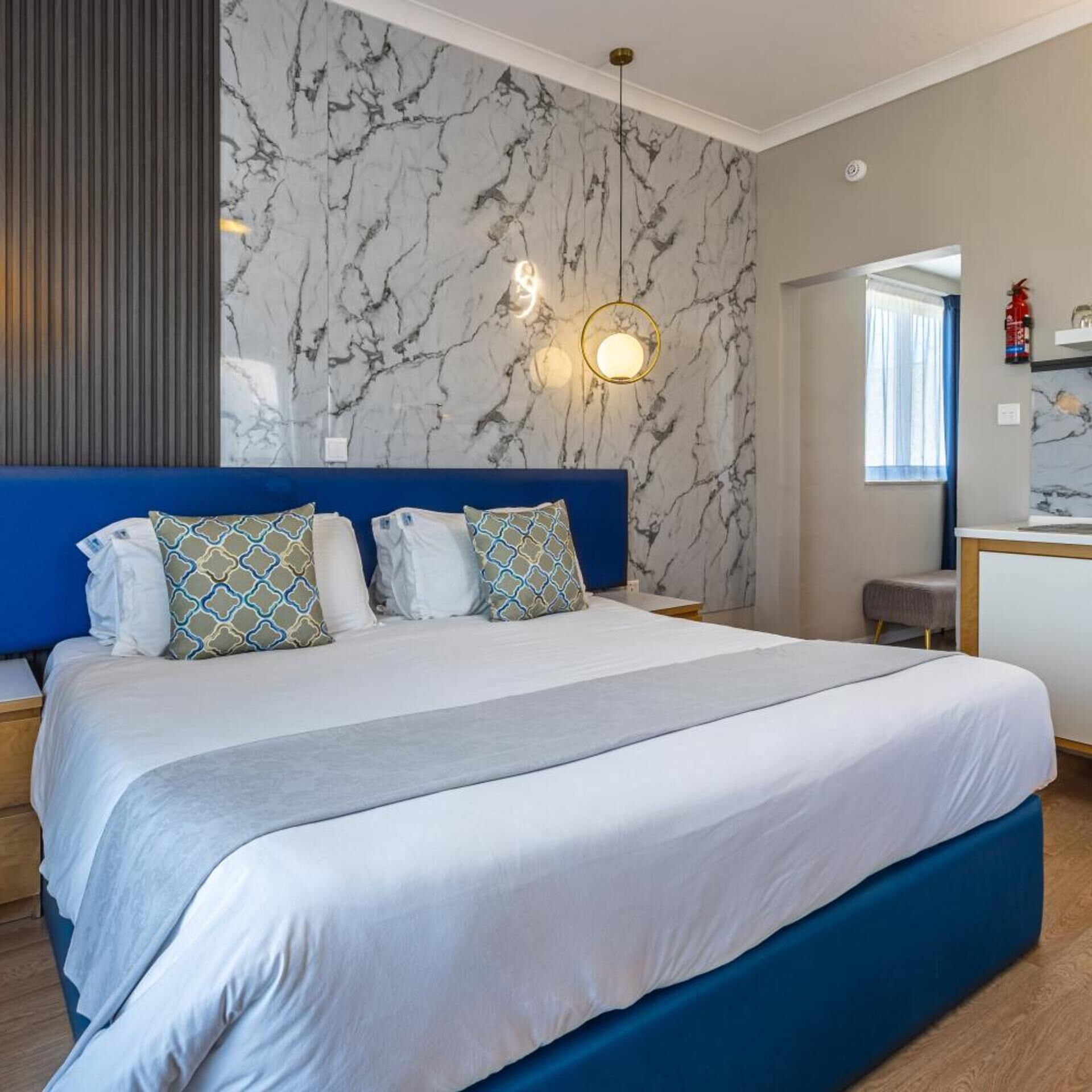 A bedroom with a marble wall, a blue headboard, a bed with white bedding and patterned pillows, a bedside table, and a hanging light fixture.