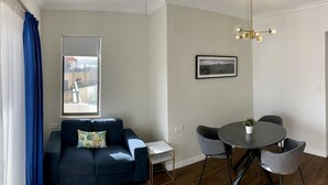 A modern living room with a dark blue sofa, a small side table, a round dining table with four chairs, a hanging light fixture, and a framed picture on the wall. - Park Lane (St. Paul's Bay)
