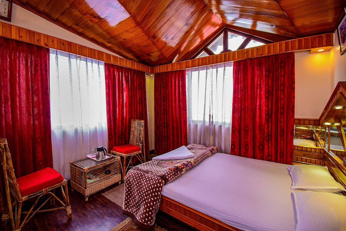 A room with a wooden ceiling, red curtains, a bed with white bedding, a wicker chair with a red cushion, and a small table with a kettle.