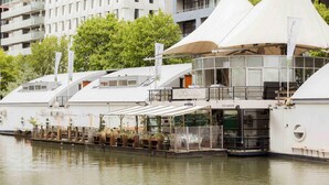A waterfront restaurant with outdoor seating, a glass facade, and a view of the river.