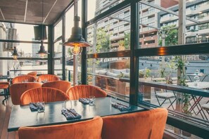 Un comedor interior con una mesa grande y sillas naranjas, ventanas amplias que ofrecen vistas a edificios y follaje exterior.