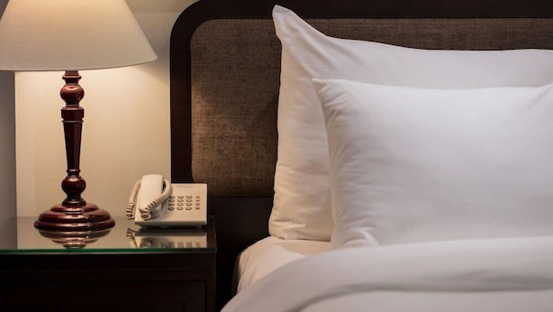 Un lit bien fait, avec du linge de lit blanc, une lampe de chevet et un téléphone posé sur une table en verre à côté du lit.