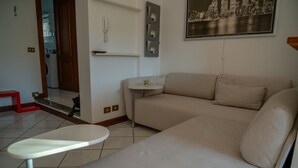 Een woonkamer met een beige bank, een klein rond tafeltje, een aan de muur bevestigde telefoon en een lijst met een stadsgezicht.