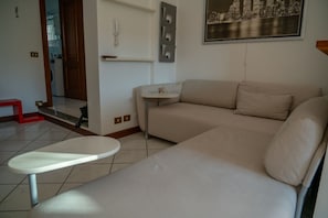 Ein Wohnzimmer mit einem beigen Sofa, einem kleinen runden Tisch, einem an der Wand montierten Telefon und einem Stadtbild in einem Bilderrahmen.