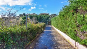 Un vialetto in pietra conduce a un edificio con tetto in tegole, immerso nel verde sotto un cielo sereno.