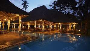 An outdoor pool area with thatched-roof structures and seating arrangements. - Neptune Village Beach Resort & Spa (Diani Beach)