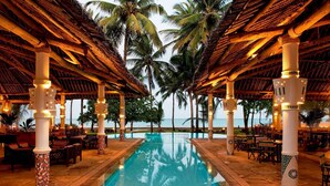 A pool area with a clear view of the ocean, surrounded by palm trees and covered with a thatched roof. - Neptune Village Beach Resort & Spa (Diani Beach)