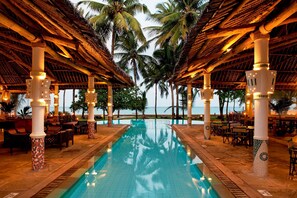 A pool area with a clear view of the ocean, surrounded by palm trees and covered with a thatched roof. - Neptune Village Beach Resort & Spa (Diani Beach)