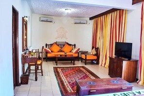 A living room with a sofa set, a television, a wooden cabinet, and a patterned rug. - Neptune Village Beach Resort & Spa (Diani Beach)