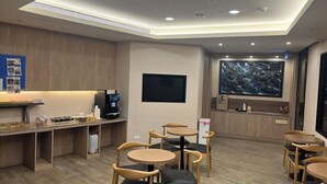 Un interior moderno con suelo de madera, mesas redondas y sillas. Hay una zona de recepción con mostrador, televisor y un cuadro grande en la pared.