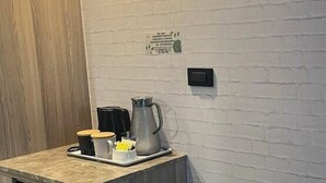 Una kitchenette compacta con encimera de madera, una cafetera y un hervidor. Hay un refrigerador pequeño y una silla metálica con bandeja.