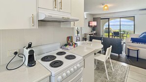 A modern kitchen with white cabinets, a built-in stove, and a dining area with a view of the sea. - Beachside Apartment Overlooking Casey’s Beach (Batehaven)