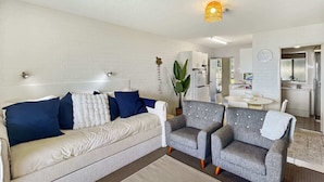 A modern living room with a sofa, two armchairs, a dining table, and a kitchen area. - Beachside Apartment Overlooking Casey’s Beach (Batehaven)
