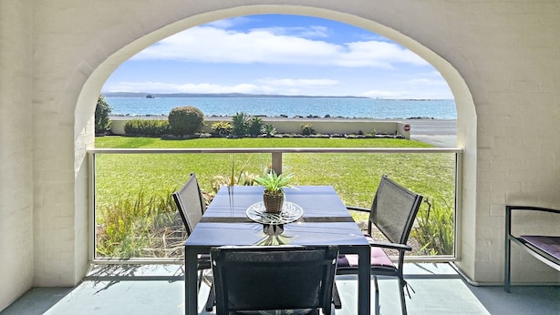 A dining area with a table and chairs, a potted plant, and a view of the sea through an arched window. - Beachside Apartment Overlooking Casey’s Beach (Batehaven)