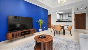 Un soggiorno moderno con un'ampia TV a schermo piatto, un tavolino in legno, una parete d'accento blu, un'area pranzo con sedie e una cucina sullo sfondo.