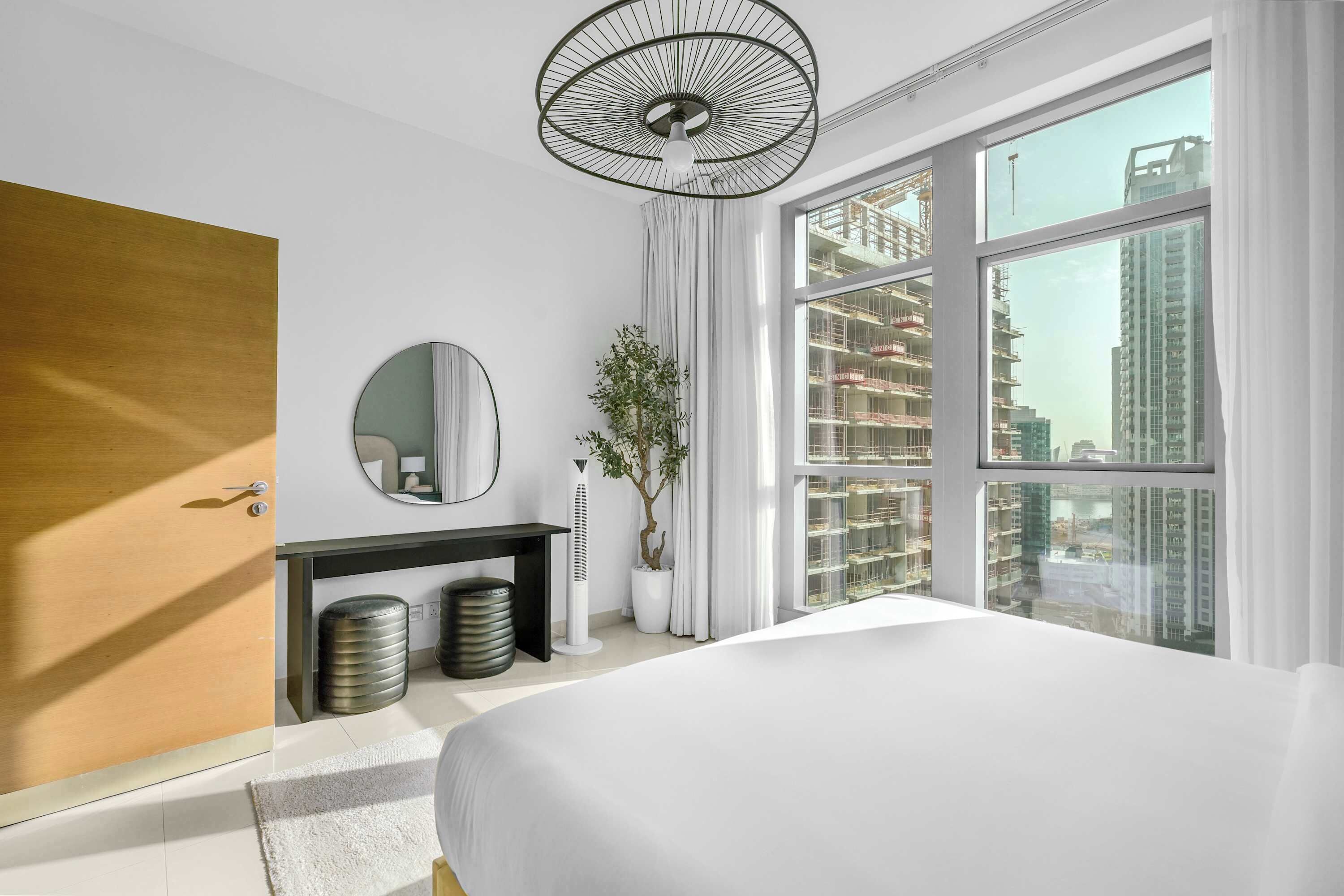 Ein modernes Hotelzimmer mit einem großen Bett, einem runden Spiegel, einer Zimmerpflanze, einem Deckenventilator und Blick auf die Stadt durch große Fenster.