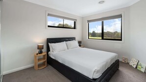 A bedroom with a large bed, two windows, a bedside table with a lamp, and a small shelf. - Family-Friendly 4-Bed Home with Courtyard (Holt)