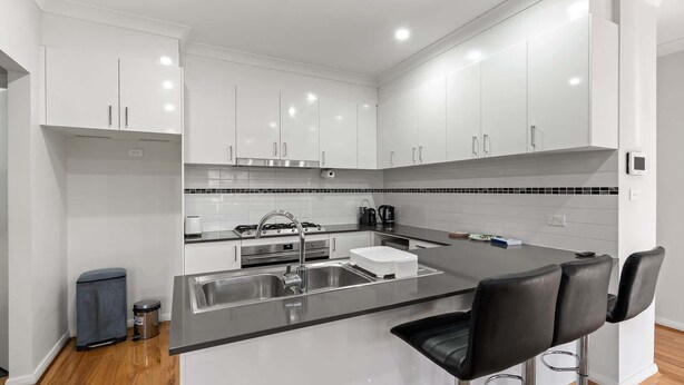 A modern kitchen with white cabinets, a black countertop, stainless steel sink, and two bar stools. - Family-Friendly 4-Bed Home with Courtyard (Holt)